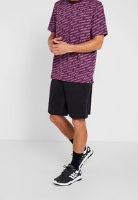 Långärmad lila skjorta med vit "UNDER ARMOUR" text, parat med svarta shorts, svartvita sneakers och svarta ankelsockor.