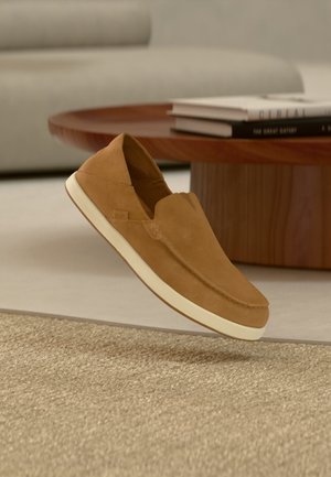 Hellbrauner Slipper mit weißer Sohle, der über einem beigen strukturierten Teppich schwebt, mit einem Holzcouchtisch und Büchern im Hintergrund.