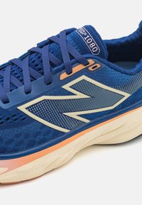 Baskets de course bleues avec une tige en tissu texturé, un logo prominent et des accents oranges. Elles possèdent une semelle rembourrée avec des rainures pour plus de flexibilité.