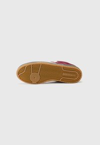 Red and white sneaker with a textured rubber sole, featuring a herringbone pattern and circular emblem on the bottom. Branding on the side.