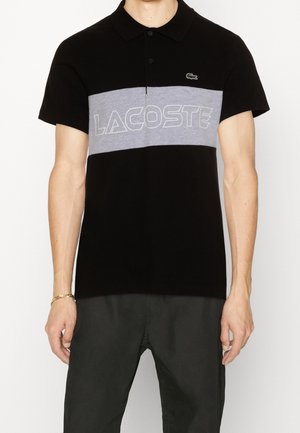 Homme portant un polo noir à manches courtes avec une bande horizontale grise et le lettrage blanc Lacoste sur la poitrine, associé à un pantalon foncé.