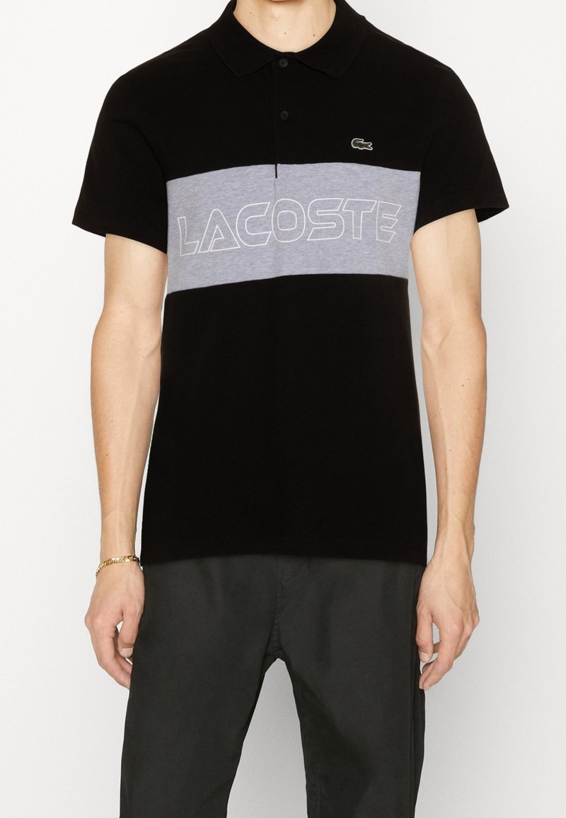 Homme portant un polo noir à manches courtes avec une bande horizontale grise et le lettrage blanc Lacoste sur la poitrine, associé à un pantalon foncé.