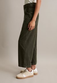 Olivgrüne weit geschnittene Hosen aus strukturiertem Stoff, mit seitlichen Taschen und Knöpfen am Saum, kombiniert mit beigen Sneakers.