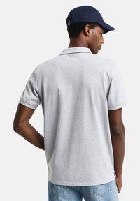 Grå polo skjorte med korte ermer, ribbestrikket krage, litt tilpasset design, sammen med en mørkeblå baseballcap, sett fra baksiden.