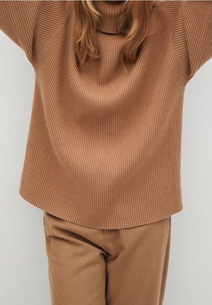 Kvinde iført en løstsiddende, ribstrikket brun sweater med høj hals og matchende brune bukser foran en ensfarvet baggrund.