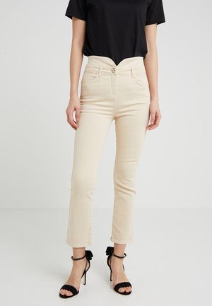 Vrouw draagt beige, slim-fit broek met hoge taille, zwart kort mouwshirt en zwarte sandalen met hoge hak, enkelsbandjes en strikken.