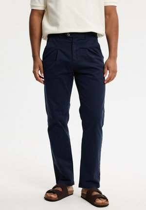 Hombre que lleva pantalones plisados azul marino, camisa beige de manga corta y sandalias marrones sobre un fondo blanco liso.