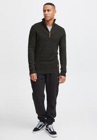 Dark green knitted pullover with a zip collar, textured fabric, ribbed cuffs, and hem. Paired with black sweatpants and black sneakers.