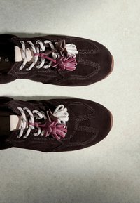 Baskets en daim marron foncé avec des lacets blancs et noirs, décorées de pampilles en cuir roses et blanches sur une surface claire.