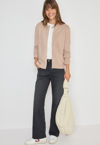 Ljus beige cardigan med ribbade muddar, bärs över en vit skjorta. Mörka utsvängda jeans och vita sneakers med röd accent. Håller en creamfärgad väska.