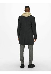 Donkergrijze jas met een lang model, gedragen over een beige hoodie. Lichtgrijze geruite broek en zwarte schoenen maken de outfit compleet.