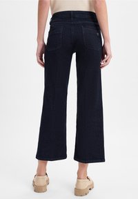 Persona vestida con jeans oscuros de pierna ancha y cortos, y zapatos beige voluminosos, mostrada desde atrás contra un fondo blanco liso.