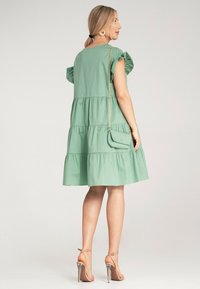 Zielona sukienka warstwowa z krótkimi falbaniastymi rękawami, okrągłym dekoltem i luźnym krojem. Uzupełniona o małą torebkę na ramię i sandały na wysokim obcasie.