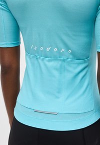 Πλάτη ενός ατόμου που φοράει μια υφασμένη τιρκουάζ ποδηλατική φανέλα με τρεις πίσω τσέπες και κείμενο "isadore" στην άνω πλάτη.