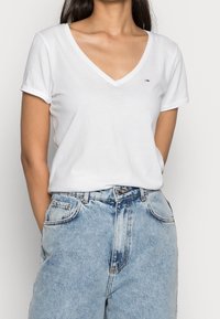 Camiseta blanca de algodón con escote en V y mangas cortas, que presenta un pequeño logo en el pecho. Combinada con jeans de mezclilla de tiro alto en azul claro.