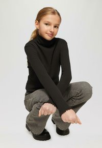 Zwarte geribbelde turtleneck met lange mouwen, grijze hoge taille jeans en zwarte chunky laarzen. Knielende houding met handen op de knieën.