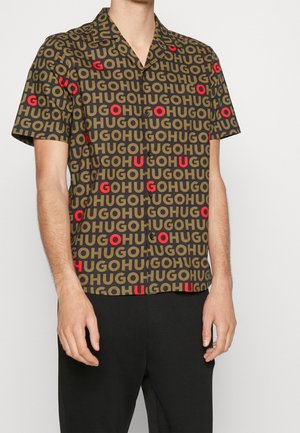 Chemise à manches courtes boutonnée avec un motif intégral de texte "HUGO" marron et rouge sur tissu noir, portée par une personne debout.