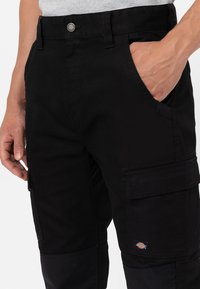 Black cargo pants made from sturdy fabric feature side pockets and a small branded patch. The waist is finished with a metal button.