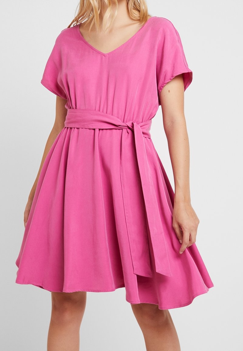 Robe courte rose avec un décolleté en V, des manches courtes et une taille nouée. Fabriquée en tissu doux, elle présente une jupe évasée et une texture lisse.