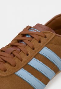 Καφέ σουέντ αθλητικό παπούτσι με ανοιχτό μπλε ρίγες της Adidas και καφέ κορδόνια που ταιριάζουν σε λευκό φόντο.