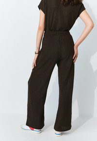 Zwarte textuur brede jumpsuit met een ontspannen pasvorm, korte mouwen en een elastische tailleband, gecombineerd met witte sneakers met rode accenten.