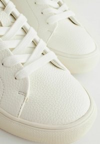 White sneakers made of textured leather with flat white laces, rounded toe, and rubber soles featuring a patterned tread.
