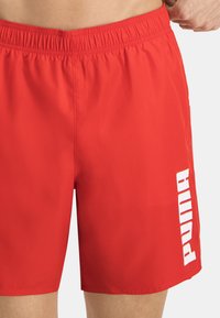 Shorts de baño rojos hechos de una tela ligera con una cinturilla elástica. Presenta un logo blanco impreso verticalmente en el lado izquierdo.