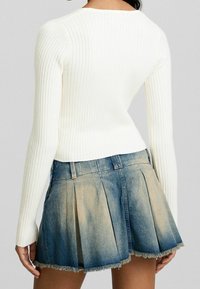 Haut blanc à manches longues côtelées, associé à une mini-jupe en denim bleu avec plis et ourlet effiloché, créant une texture en couches.