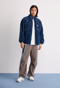 Navy fleece jacket with white stitching details, zippered front, and elastic cuffs, paired with beige cargo pants and white sneakers.