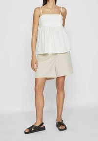 Vit ärmlös topp med elastisk midja, kombinerad med beige shorts. Modellen bär svarta sandaler. Enkel och avslappnad passform.