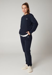 Sada navy modrého svetru a tepláků vyrobená z měkké tkaniny, s uvolněným střihem a akcentem s logem. Tenisky doplňují tento outfit.