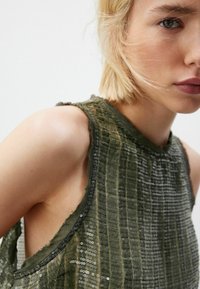 Khaki groene mouwloze top van textuurstof met pailletten, met een hoge halslijn en ruwe randen rond de armsgaten.