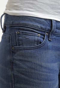 Tmavě modré džíny z denimu s malou přední kapsou, kovovými komponenty a jemnými variacemi textury v tkanině.