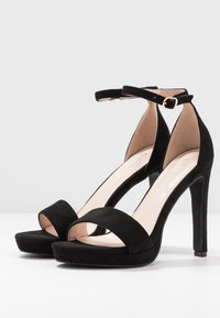 Svarta mocka högklackade sandaler med en tjock rem vid tån, ankelrem med ett guldspänne och en plattform för extra höjd.