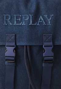 Denim nahrbtnik v temno modri barvi z vtisnjenim logotipom "REPLAY". Ima trdne sponke, zapiranje na vrvico in teksturirane tkanine.