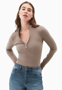 Kvinde med langt brunt hår iført taupe ribbet langærmet top, udrullet ved kraven, og højtsiddende blå jeans, med hænderne hvilende på hofterne.