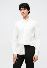 Camicia bianca a maniche lunghe con bottoni, taschino sul petto e colletto standard, abbinata a pantaloni neri su uno sfondo neutro.