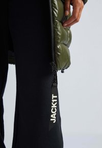 Olivově zelená péřová bunda s lesklou, prošívanou strukturou; má černý zip a štítek s bílým textem "JACK1T." Černé kalhoty viditelné pod ní.