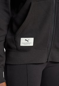 Sort lynlås hættetrøje med tekstureret stof, der har et hvidt mærke med "PUMA ATHLETICS" logoet på forsiden. Slim fit design.
