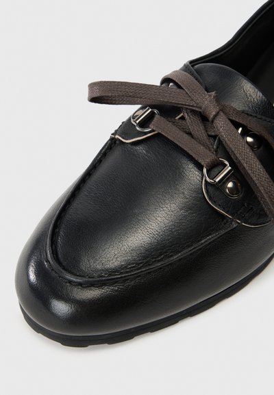 Svarta läderloafers med brunt texturerat snöre som är bundet i en rosett över silverfärgade öglor mot en ren vit bakgrund.