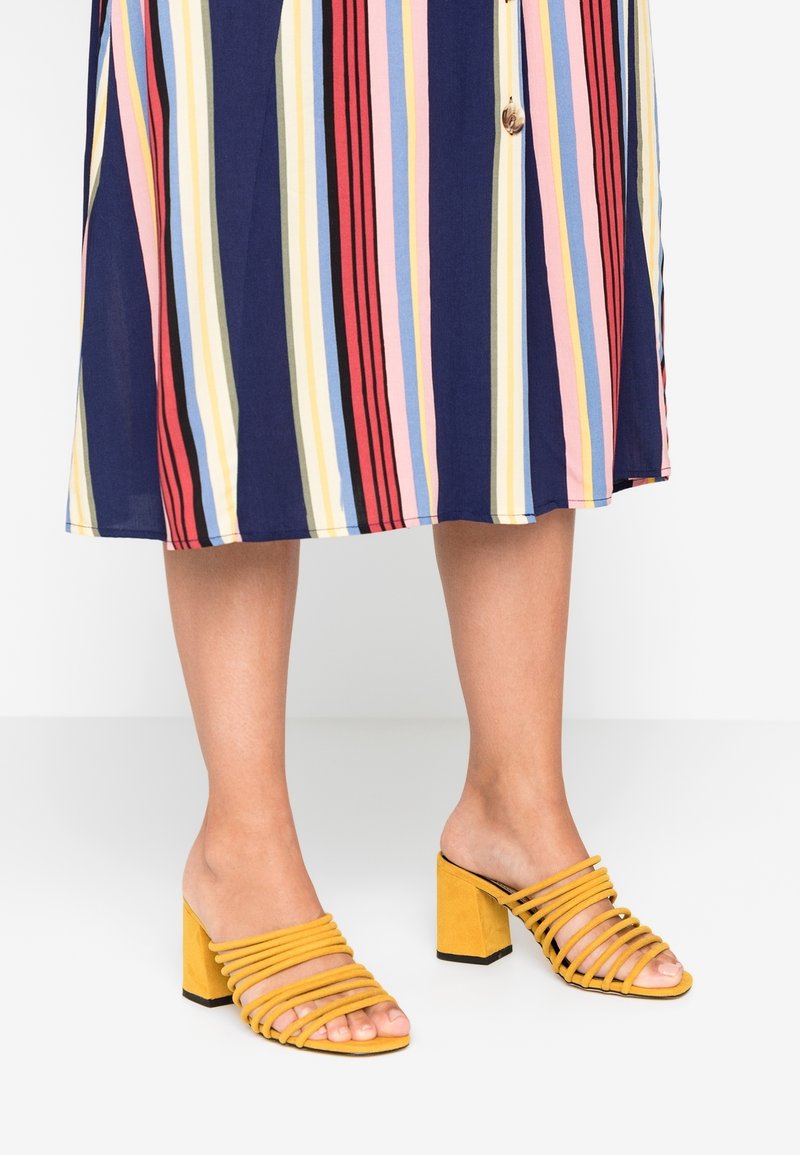 Keltaiset mokkanahkaisten liukusandaalit, joissa on monivärinen raidoitus, leveät vaakasiteet, avoin kärki ja paksu musta kantapää.