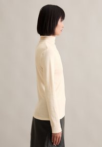 Cremefarbener Rollkragenpullover mit langen Ärmeln, körperbetontes Design, weiche Textur, glattes Material. Kombiniert mit einem dunklen Lederrock, seitliche Ansicht.