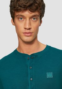Tirkizna pamučna henley majica s okruglim izrezom, tri gumba sprijeda i mali izvezeni logo na lijevoj strani prsnog koša.
