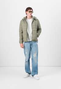 Olivengrønn pufferjakke med hette, lysegrå t-skjorte, slitte lyseblå jeans og hvite joggesko. Solbriller med klare glass.