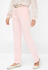 Lichtroze broek met een rechte snit, voorzien van een hoge taille en een riem. Gecombineerd met beige platte schoenen, tegen een witte achtergrond.
