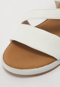 Sandale en cuir blanc avec deux larges brides croisées. La semelle intérieure est beige, avec une texture douce, et la semelle est plate et légère.