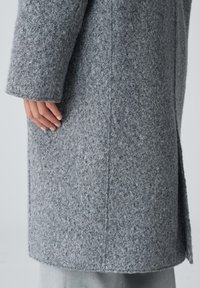 Grauer Wollmantel mit strukturiertem Finish, gerade geschnitten und mit seitlichem Schlitz. Eine Nahaufnahme zeigt die flauschige Oberfläche des Stoffes und die Nähdetails.