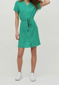Grön klänning med vita prickar, korta ärmar, knäppfront och justerbart knyt vid midjan; kombinerad med vita sneakers.
