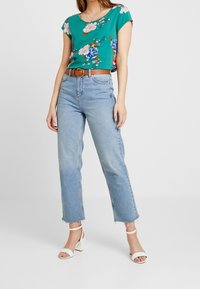 Květinový zelený top s krátkým rukávem, světle modré džíny s vysokým pasem, hnědý opasek a bílé sandály na blokovém podpatku, vystavené na osobě, která stojí.