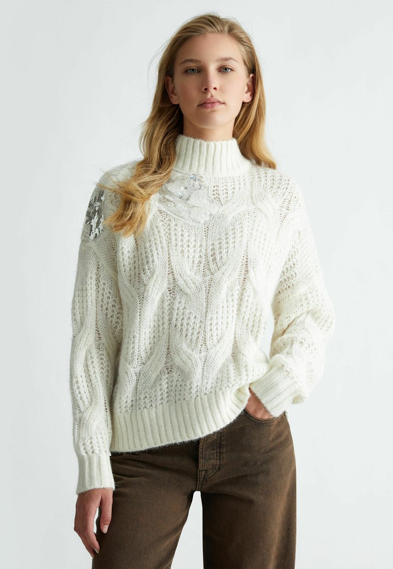 Maglione bianco lavorato a maglia con colletto alto, caratterizzato da un motivo a trecce e dettagli in paillettes sulla spalla sinistra. Polsini e orlo a coste.